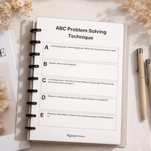 May include: A notebook with the text "ABC Problem Solving Technique" and sections labeled A through E. The notebook is open, with pens and paperclips nearby. The background includes dried flowers and a magazine.