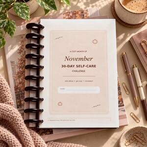 Può includere: Una pagina di agenda rilegata a spirale intitolata A Cozy Month of November 30-Day Self-Care Challenge. La pagina presenta uno sfondo beige chiaro con piccole icone di zucche e foglie, il testo slow down, get cozy, reconnect e una riga per un nome.