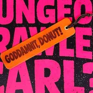 Könnte beinhalten: Ein rechteckiges orangefarbenes Lesezeichen mit der dunkelbraun eingravierten Aufschrift "GODDAMNIT, DONUT!", einem Loch an der Oberseite und einer braunen Lederschnur. Das Lesezeichen liegt auf einem schwarzen Hintergrund mit großen, pinken Buchstaben.
