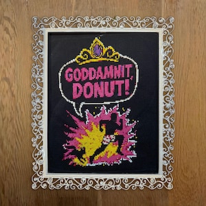 May include: Cross-stitch art in a silver ornate frame. The black background features the text "GODDAMNIT, DONUT!" in a speech bubble, with a crown above. A running figure is silhouetted against a pink and yellow explosion.