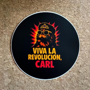 May include: Circular black sticker with a cat illustration wearing sunglasses and a crown. The text "VIVA LA REVOLUCIÓN, CARL" is in yellow and red. The cat has orange and red accents, creating a fiery effect.