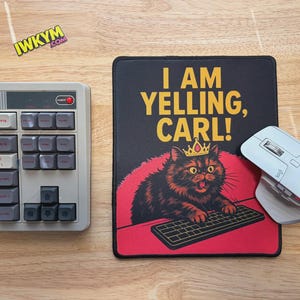Può includere: Un'impostazione del computer con una tastiera retrò, un mouse wireless bianco e un tappetino per mouse nero. Il tappetino per mouse presenta il testo "I AM YELLING, CARL!" e un gatto dei cartoni animati con una corona.