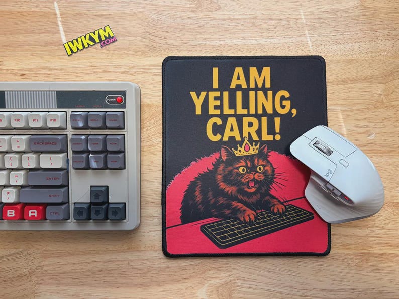 Op de afbeelding: Een computeropstelling met een retro toetsenbord, een witte draadloze muis en een zwarte muismat. De muismat heeft de tekst "I AM YELLING, CARL!" en een cartoonkat met een kroon.