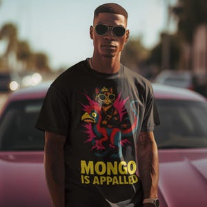 Può includere: T-shirt nero con una grafica di un gatto con occhiali da sole e una corona, in sella a una creatura simile a un uccello. Il testo "MONGO IS APPALLED" è in giallo. La persona indossa occhiali da sole e un orologio.