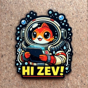 Pu&ograve; includere: Un adesivo con un pesce rosso dei cartoni animati in tuta spaziale, che tiene in mano un tablet. Il pesce rosso &egrave; arancione e bianco, con grandi occhi e un'espressione sorridente. Il testo "HI ZEV!" &egrave; giallo.