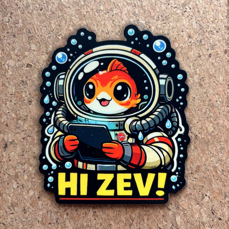 Pu&ograve; includere: Un adesivo con un pesce rosso dei cartoni animati in tuta spaziale, che tiene in mano un tablet. Il pesce rosso &egrave; arancione e bianco, con grandi occhi e un'espressione sorridente. Il testo "HI ZEV!" &egrave; giallo.