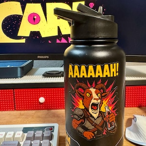 May include: Black water bottle with a black flip-top lid. The bottle features a sticker with a cartoon goat character yelling and holding a bottle. The text "AAAAAAH!" is above the goat.
