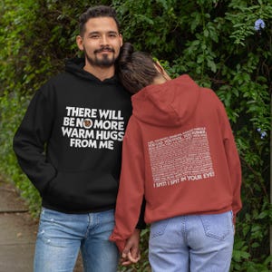 May include: Two hooded sweatshirts, one black and one red. The black hoodie features the text "THERE WILL BE NO MORE WARM HUGS FROM ME" in white. The red hoodie has text on the back that includes "I SPIT! I SPIT IN YOUR EYE!"
