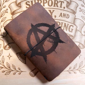 May include: A brown leather journal featuring a black anarchy symbol printed on the front cover. The journal is secured with a thin black elastic band that wraps vertically around the centre of the cover, holding the leather notebook closed.