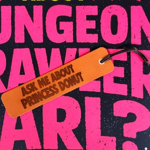 Könnte beinhalten: Ein rechteckiges Lesezeichen aus hellbraunem Leder mit dem lasergravierten Text ASK ME ABOUT PRINCESS DONUT. Das Lesezeichen hat oben eine messingfarbene Metallöse, durch die ein dünnes, dunkelbraunes Lederband als Abschluss gezogen ist.