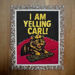 May include: Cross-stitch art piece with the text "I AM YELLING CARL!" in yellow letters on a black background. The image features a cat wearing a crown, sitting on a red surface, and a keyboard. The piece is framed with a decorative white border.