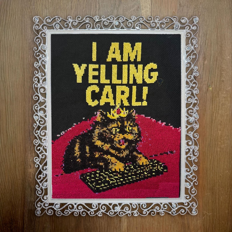 May include: Cross-stitch art piece with the text "I AM YELLING CARL!" in yellow letters on a black background. The image features a cat wearing a crown, sitting on a red surface, and a keyboard. The piece is framed with a decorative white border.