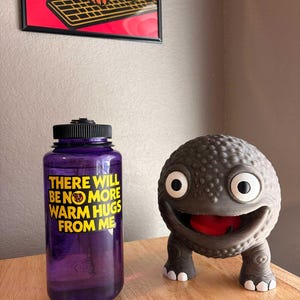 以下が含まれることがあります： 黒い蓋と「THERE WILL BE NO MORE WARM HUGS FROM ME」の黄色い文字が入った紫色のウォーターボトル。その隣には、大きな白い目と赤い口を持つ、灰色の笑顔の生き物がいます。背景には、猫のアート作品が額縁に入って飾られています。