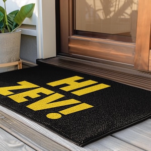 Puede incluir: Felpudo rectangular negro con el texto "HI ZEV!" en amarillo. La alfombra está colocada en un porche de madera frente a una puerta. Una planta en maceta es visible en el fondo.