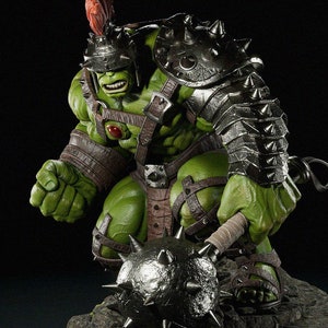 Puede incluir: Una figura verde de un personaje musculoso con armadura de batalla, sosteniendo una maza con púas. La figura tiene un casco con una pluma roja y está posada sobre una base de piedra. La armadura es plateada y marrón, con correas de cuero.