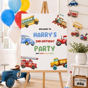 May include: A white rectangular birthday party sign on a wooden easel featuring watercolour illustrations of a yellow school bus, a blue car, a red tractor, a red fire engine, and a yellow dump truck with the text Welcome to Harry's 2nd Birthday Party.