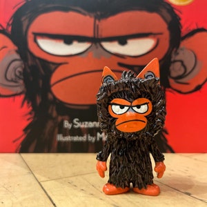 May include: A figurine of a grumpy monkey with orange hands and feet stands in front of a book titled "Grumpy Monkey." The book cover is red with a large illustration of a grumpy monkey face. The figurine has a brown body and orange accents.