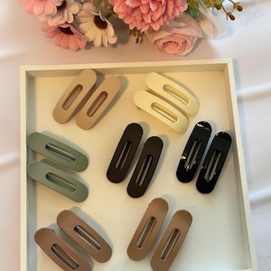 May include: A collection of ten oval-shaped hair clips in various matte and glossy finishes, including shades of beige, sage green, cream, dark brown, and black, arranged in pairs on a white tray with pink and white artificial flowers above.