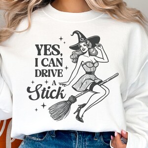 May include: White sweatshirt with a black and white graphic of a witch riding a broomstick. The text on the sweatshirt reads 'YES, I CAN DRIVE A STICK'.