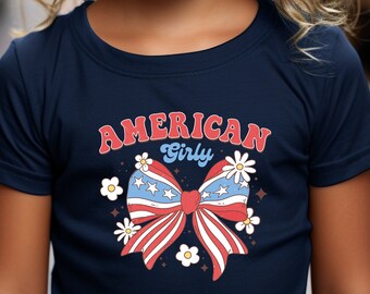 May include: A young girl with curly blonde hair is wearing a navy blue t-shirt with a red, white, and blue bow with stars and the text "American Girly".