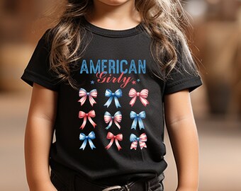 May include: A young person wearing a black t-shirt with the text "AMERICAN Girly" and a pattern of red, white, and blue bows. They are also wearing a black cowboy hat and black jeans.