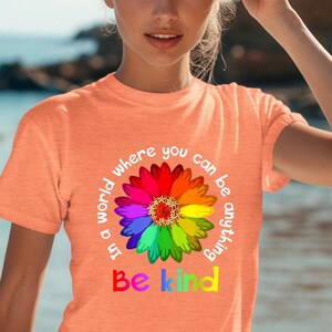Könnte beinhalten: Ein pfirsichfarbenes T-Shirt mit einem Regenbogenblumen-Motiv und dem Text "In einer Welt, in der du alles sein kannst, sei freundlich".