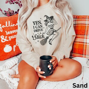 May include: A beige sweatshirt with a black and white illustration of a witch riding a broomstick. The text on the sweatshirt reads "Yes, I can drive a stick".