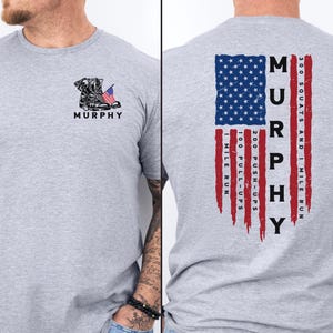 May include: A light gray t-shirt featuring a black and white graphic of a combat boot with an American flag and the name "MURPHY". The back of the shirt displays a distressed American flag design with fitness goals.