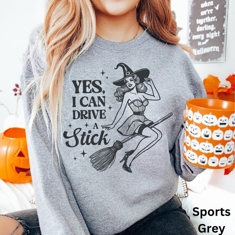 May include: A grey sweatshirt featuring a graphic of a witch flying on a broomstick. The text on the sweatshirt reads "YES, I CAN DRIVE A STICK".