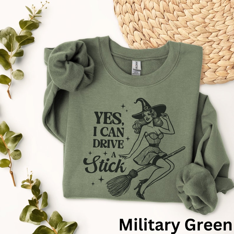 May include: A green sweatshirt featuring a witch riding a broomstick with the text "YES, I CAN DRIVE A STICK".