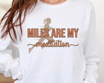 May include: White long-sleeve shirt with a brown silhouette of a woman running and the text "MILES ARE MY meditation" in orange.