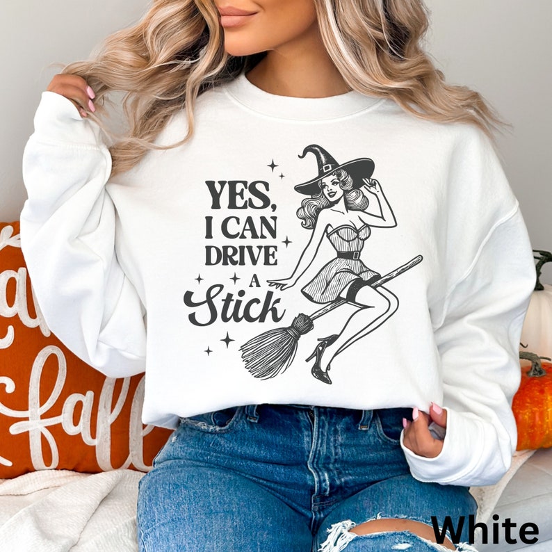 May include: White sweatshirt with a black and white graphic of a witch riding a broomstick. The text on the sweatshirt reads 'YES, I CAN DRIVE A STICK'.