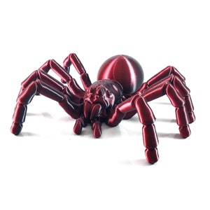 Peut inclure: Une araignée rouge imprimée en 3D avec une finition métallique brillante. L'araignée a huit pattes et un corps rond et volumineux.