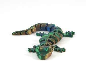 May include: A 3D printed articulated gecko figurine with a green, blue, and gold iridescent finish. The gecko is posed in a walking position with its head tilted down.