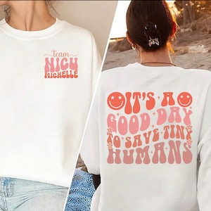 May include: White crew neck sweatshirt with the text "Team NICU Michelle" on the front and "It's a good day to save tiny humans" on the back. The text is in a retro font with pink and orange colors.