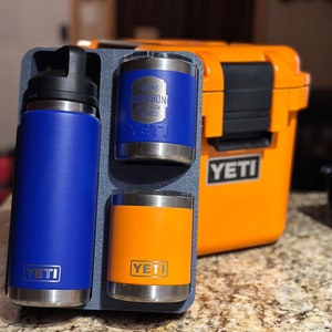 May include: A set of three YETI brand drinkware items. A blue YETI Rambler 26 oz bottle, a blue YETI Rambler 10 oz cup with the text "I drink bourbon and know things", and an orange YETI Rambler 10 oz cup. The items are displayed in a gray and black carrying case.