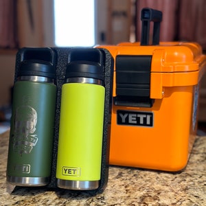 Op de afbeelding: Twee YETI-merk roestvrijstalen waterflessen, een groene met een schedelgrafiek en de tekst "The Way To Go", en een gele, in een zwarte draagtas naast een oranje YETI-koeler.