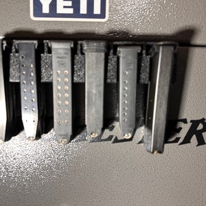 May include: A collection of six metal magazines, each with a different design, are mounted on a gray surface. The magazines are of varying lengths and widths, with some featuring visible bullet indicator holes. The image is taken indoors.