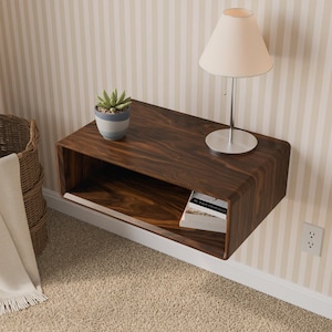 May include: A dark brown wood floating nightstand with a rectangular open shelf design. A small potted succulent and a table lamp with a beige conical shade sit on the top surface. A white book is placed inside the open storage compartment.