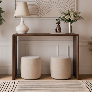 May include: A modern entryway with a wooden console table, two beige ottomans, a white lamp with a cream shade, a black and white geometric print, a vase with white flowers, a wooden sculpture, and a potted plant.