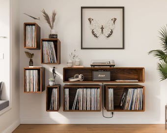 Op de afbeelding: Een aan de muur gemonteerde platenspelerplank met een platenspeler, koptelefoon en een kleine versterker. De plank is gemaakt van donker hout en heeft een modern ontwerp. Er is een vlinderprint op de muur boven de plank.