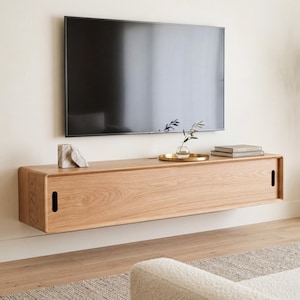 May include: A wall-mounted light oak media console featuring two sliding doors with vertical cutout handles. The rectangular wooden unit is positioned beneath a flat-screen television and holds a small stone sculpture, a gold tray, and books.