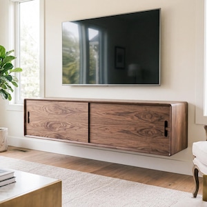 May include: A wall-mounted walnut wood media console featuring two sliding doors with vertical cutout handles, positioned directly beneath a large flat-screen television on a light-colored wall in a bright living room setting.