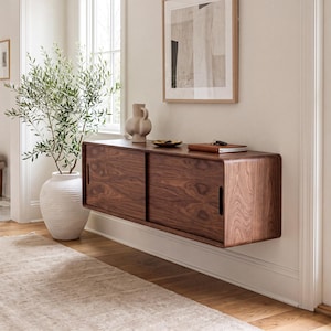 May include: A wall-mounted dark wood cabinet with rounded edges and two sliding doors featuring vertical handle cutouts. The cabinet is positioned horizontally on a light-colored wall, providing a sleek storage solution for a home entryway.