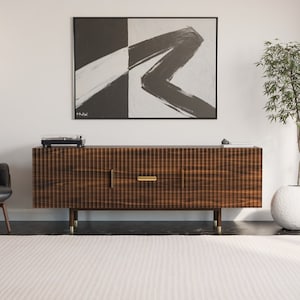 May include: A mid-century modern living room with a black leather armchair, a wooden cabinet with gold hardware, a black and white abstract painting, a potted plant, and a white rug.