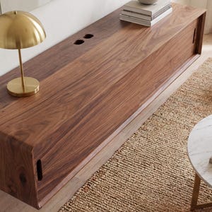 K&ouml;nnte beinhalten: Eine lange, rechteckige, wandmontierte Medienkonsole aus dunkelbraunem Holz mit einer glatten, durchgehenden Maserung. Sie verf&uuml;gt &uuml;ber Schiebet&uuml;ren mit vertikalen Griffaussparungen und zwei kreisf&ouml;rmige Kabeldurchf&uuml;hrungen auf der Oberseite.