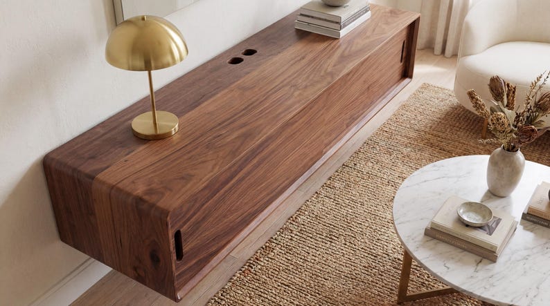 K&ouml;nnte beinhalten: Eine lange, rechteckige, wandmontierte Medienkonsole aus dunkelbraunem Holz mit einer glatten, durchgehenden Maserung. Sie verf&uuml;gt &uuml;ber Schiebet&uuml;ren mit vertikalen Griffaussparungen und zwei kreisf&ouml;rmige Kabeldurchf&uuml;hrungen auf der Oberseite.