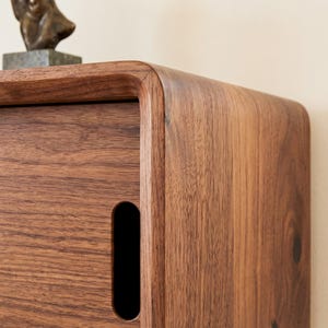 K&ouml;nnte beinhalten: Ein dunkelbrauner Holzschrank mit abgerundeten Ecken und einem vertikalen, ovalen Griffausschnitt an der Frontplatte. Die Holzmaserung ist auf der gesamten Oberfl&auml;che sichtbar und unterstreicht die nat&uuml;rliche Textur und die satten Farbt&ouml;ne.