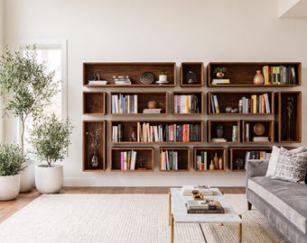 May include: A collection of rectangular dark wood wall-mounted shelves arranged in a grid pattern, filled with various books, small decorative objects, and vases. The shelves are mounted on a plain white wall above a gray sofa and a marble coffee table.