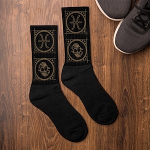 Könnte beinhalten: Schwarze Socken mit goldenen Sternzeichen-Fischen. Jede Socke hat zwei Fisch-Symbole, eines oben und eines in Knöchelhöhe. Die Socken liegen auf einer Holzoberfläche, im Hintergrund ein Paar graue Turnschuhe.